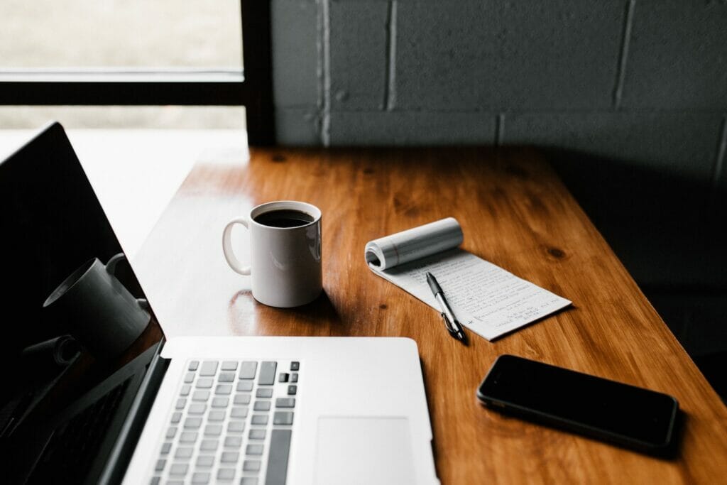 Image of a modern, wooden desk. On it sits a laptop computer, coffee mug, notebook and pen, and glasses case.